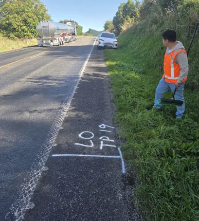 A TTAP engineer in high-visibility clothing marking a test-pit location on a state-highway shoulder, with a tanker and traffic passing in the live lane.