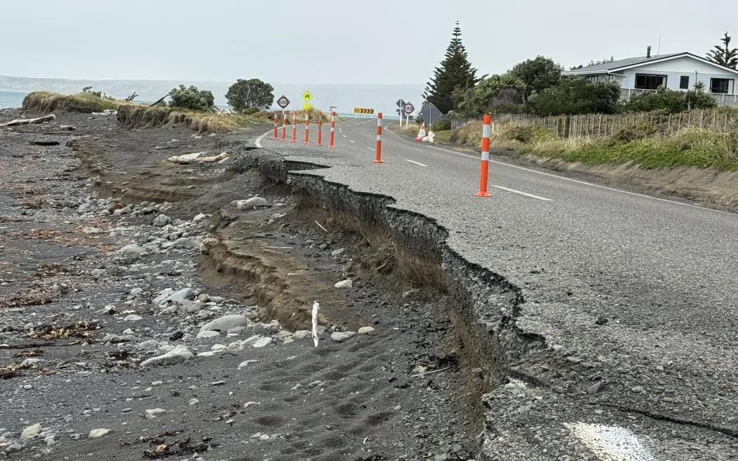 Cape Palliser Road with the seaward shoulder washed out — the carriageway narrowed to a single lane, coned off, with the sea breaking against the exposed edge and houses immediately inland.