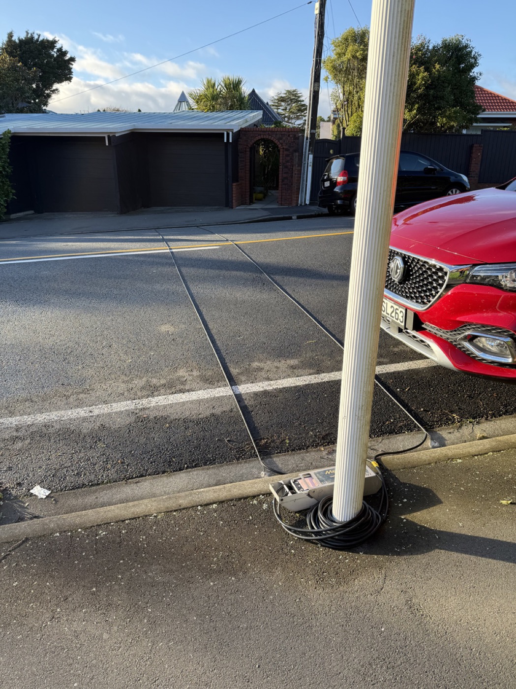 Pneumatic tube counter cables strapped to a power pole on a quiet New Zealand residential street, with houses and a passing car visible.